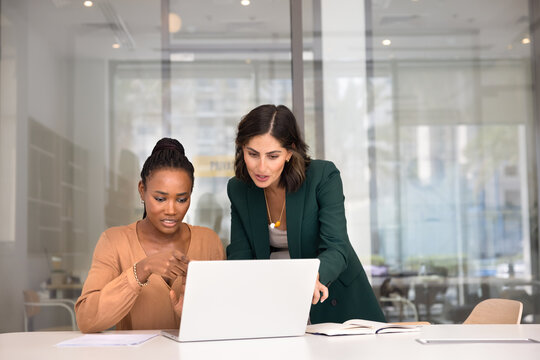 Two multiethnic female coworkers working together at laptop, pointing at screen, discussing task. Young Black professional asking colleague for advice, feedback, showing content on computer