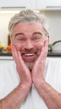 Middle-aged man at home hears positive news, grabs cheeks says wow smiles wide eyes shine with joy. Mature guy on sofa laughs claps hands breath quick heart lifts relief pure surprise and bright mood.