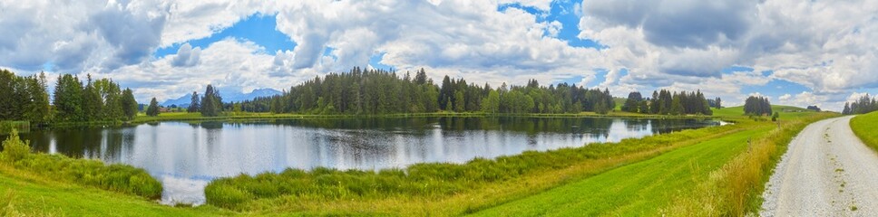 Grosses Panorama in der Nähe der Ortschaft “Seeg im Allgäu“ mit einem Teich im Vordergrund.