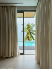 View from room of luxury villa at sunny day, sun loungers for sunbathing, umbrellas from the sun, pillows for swimming in the pool, palm tree on background