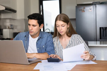 Young couple sits in modern kitchen reviewing financial documents, using laptop and calculator, involved in managing household expenses, mortgage payments, investment planning. Responsibility, budget