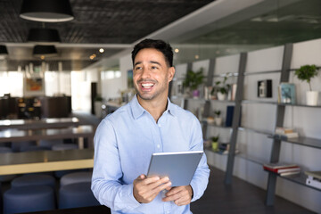 Happy young Hispanic businessman holding digital tablet, standing in office co-working space, looking away, smiling, laughing, using device for work communication, online business management