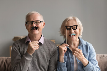 Playful timeless love. Portrait overjoyed energetic older husband wife retirees sit on cozy couch looking at camera with fake moustache and funny paper glasses on sticks for comic family photo session