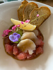 Beautifully decorated tuna tartare dish with avocado and scallop slices, lemon, bread chips and decorated with flowers on a white plate