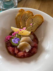 Beautifully decorated tuna tartare dish with avocado and scallop slices, lemon, bread chips and decorated with flowers on a white plate