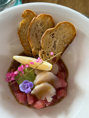 Beautifully decorated tuna tartare dish with avocado and scallop slices, lemon, bread chips and decorated with flowers on a white plate