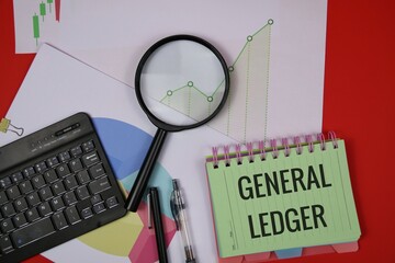 A notebook labeled "General Ledger" sits among charts, a magnifying glass, a keyboard, and pens, suggesting financial analysis, bookkeeping, and data review. isolated on a red surface.