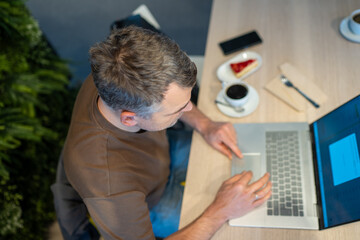 Overhead view of a man using a laptop at a table with coffee and a slice of cake filmed from the back with no face visible