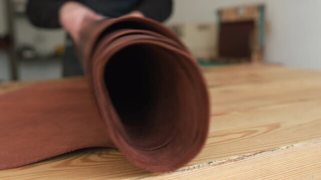 Leather crafter unrolling brown hide on wooden workbench in studio workshop, preparing material for handmade goods