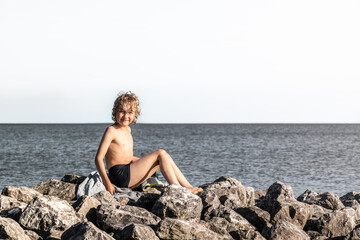A young boy sits on a towel placed on rocks by the water. He smiles at the camera on a sunny summer day while wearing swim trunks and enjoying the view.
