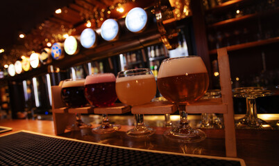 A selection of craft beers in tasting glasses placed on a wooden rack at a bar counter. Warm pub lighting and cozy atmosphere.