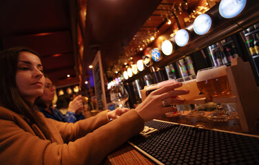 Two women tasting different craft beers at a bar counter in a cozy pub. Beer flight on wooden holders, warm interior lighting, social lifestyle atmosphere.
