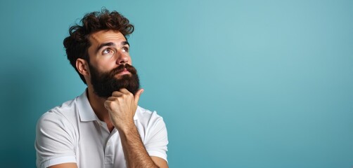 Bearded man wears polo shirt. He rests chin on hand, looking up and away, lost in deep thought and contemplation. Blue backdrop for ideas, questions, or problem solving.
