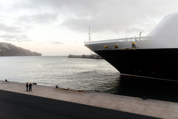 Two individuals standing on a dock near a large luxury cruise ship, with calm waters and distant hills creating a serene maritime atmosphere at sunset