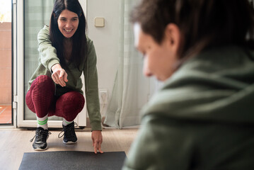 Two friends are working out together at home in a bright, relaxed environment. One of them smiles while guiding and encouraging the other, creating a natural scene that conveys support, motivation, an