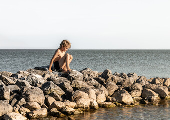 A young boy crawls on boulders near the water at the beach. He wears swimming trunks and enjoys the warm summer day by the seaside.
