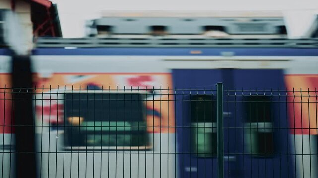 Blurred high speed train with colorful graphics rushes past behind a metal fence, conveying motion, travel and urban transportation infrastructure