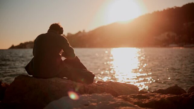 A man sitting alone on coastal rocks, looking down as the sun sets over the sea and hills, creating a warm contemplative mood