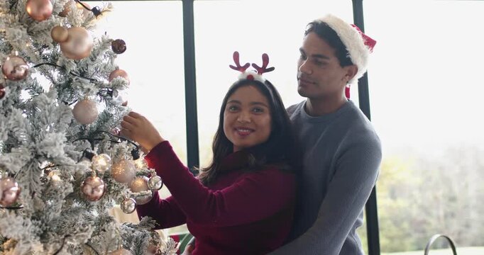 Romantic couple decorating Christmas tree selecting and hanging bauble beside window, copy space