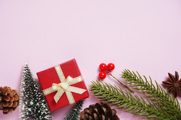 Christmas flat lay on a pink background featuring a red gift box with gold ribbon, pine branches, red berries, and miniature snow-covered Christmas trees. 