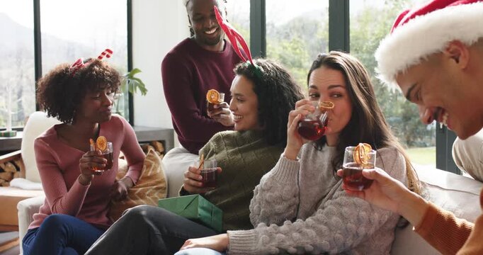 Diverse friends raising mugs after friend offering mulled drink, unwrapping gift in living room