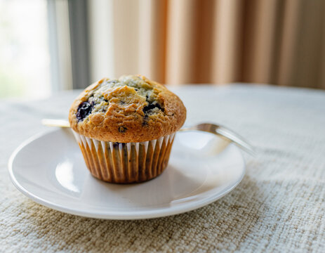 Delicious freshly baked blueberry muffin served on a white plate with cutlery by a sunlit window perfect for a cozy morning breakfast or dessert treat. - Powered by Adobe