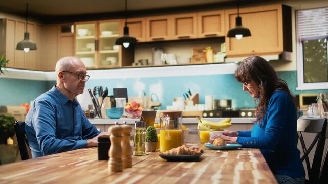 Senior male pensioner browsing webpages on laptop and wife showing him the timeout symbol, getting angry at his retirement hobbies. Absent husband ignoring woman during breakfast. Camera A.