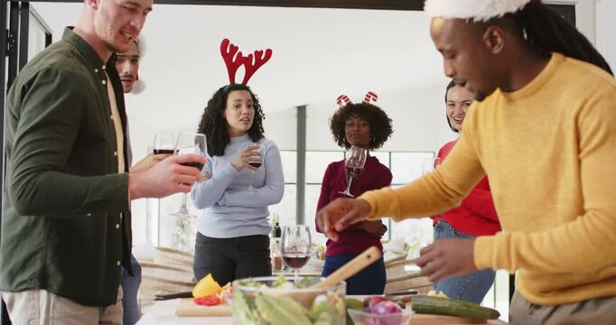 Diverse friends wearing Santa hats holding glasses, urging Black man slicing cucumber tossing salad