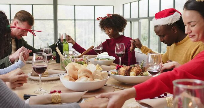 Diverse friends holding hands praying in dining room, camera descending focusing roast chicken