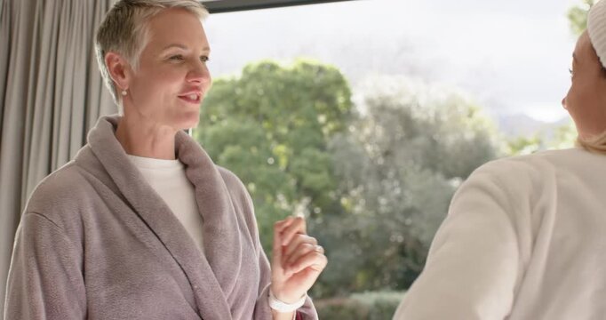 Female friends standing by glass door, one in lavender robe smiling, greeting companion sharing joy