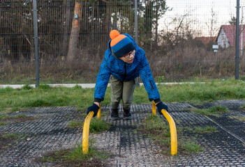 Boy in blue jacket doing push-ups on low parallel bars at outdoor workout area with rubber mat surface on cold autumn day. Concept of street calisthenics, youth fitness training and healthy lifestyle.
