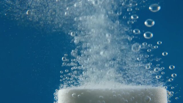 Effervescent tablet dissolving in water, creating bubbles and fizzing motion against a blue background, close-up shot