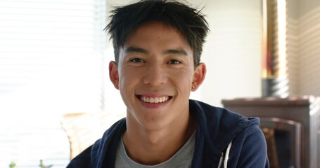 Asian man sitting facing camera in living room with blinds, shifting to smiling and nodding