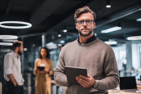 A confident male professional holds a digital tablet in a modern office looking at the camera - Powered by Adobe
