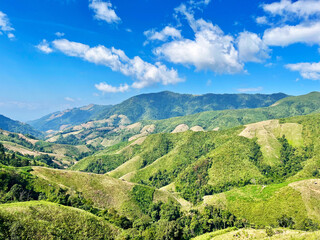 mountain landscape with blue sky, A wide mountain landscape under a bright blue sky, showcasing rolling green hills, distant ridges, soft sunlight that highlights the natural contours of the valley