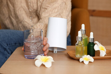 Woman with plumeria flowers and essential oils pouring water into air humidifier on table in room, closeup