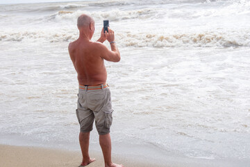 shirtless man standing on sandy beach, taking photo crashing waves with smartphone, barefoot in shallow water, tourist capturing ocean scenery during beach vacation, active retirement lifestyle