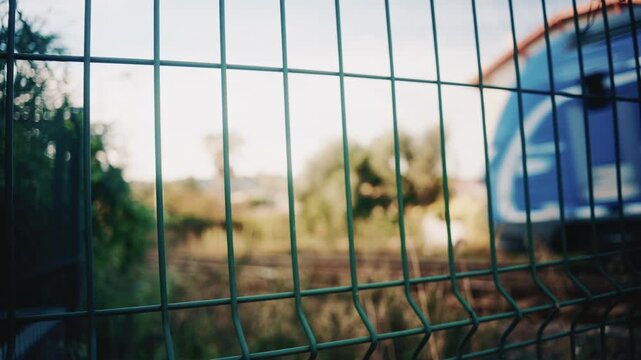 Blurry train passes behind a green metal fence, creating a layered perspective focused on the fence in the foreground