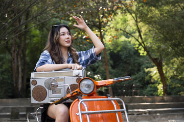Young woman sitting on orange scooter holding vintage boombox, enjoying sunny day outdoors in green park with relaxed expression