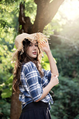 Young woman in plaid shirt and straw hat posing outdoors in sunlight with natural green background, expressing calm and relaxed mood