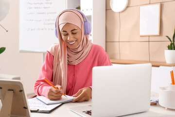 Young Muslim woman in headphones learning Arabic language online at office