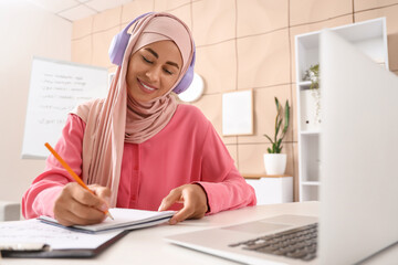 Young Muslim woman in headphones learning Arabic language online at office