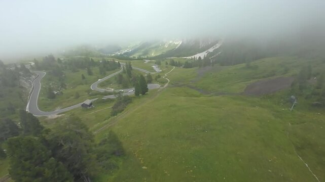 dolomites winding mountain path veiled in low cloud and mist. cinematic drone pans reveal switchback road, grassy slopes, scattered trees and soft horizon, evoking journey, exploration