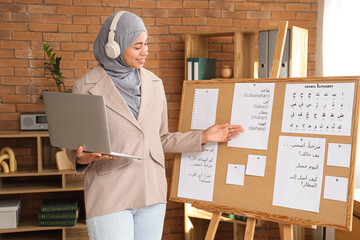 Young Muslim woman with laptop learning Arabic language online near pin board in office