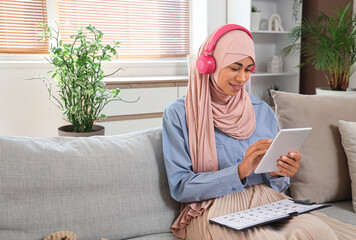 Young Muslim woman in headphones with tablet computer learning Arabic language online on sofa at home