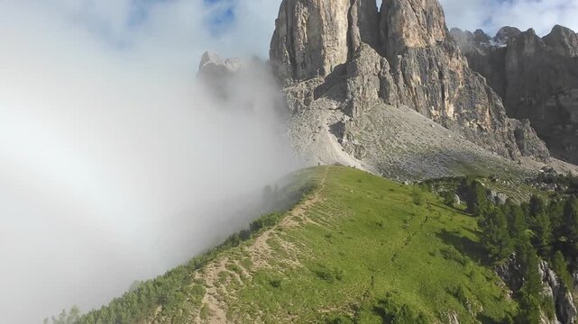 dolomites rocky ridge shrouded in mist, faint rainbow. cinematic drone glide reveals steep limestone cliffs, lush green meadow, scattered boulders and alpine pines emerging from low cloud,