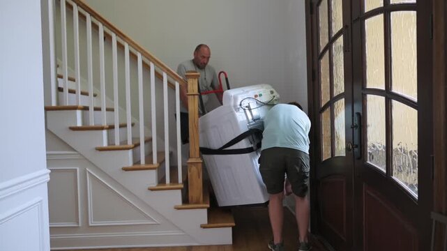 workers secure load on staircase landing, individuals carefully maneuver appliance with tools for safety, group collaboratively stabilizes and transports washing machine down staircase safely