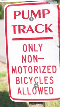 Verti al screen closeup pump track sign Johnson City park, red and white metal notice stating only non-motorized bicycles allowed, weathered bolt and post, grassy background, daytime