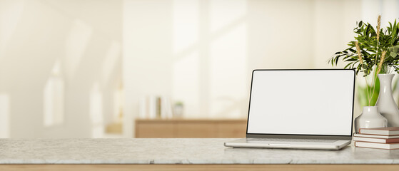 Blank screen laptop and books aside plant vase on marble counter table in a white room with sunlight