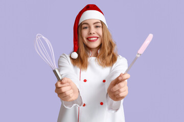 Young female chef in Santa hat with whisk and spatula on purple background